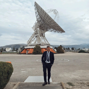 Un uomo in piedi, in giacca e cravatta, sullo sfondo un satellite