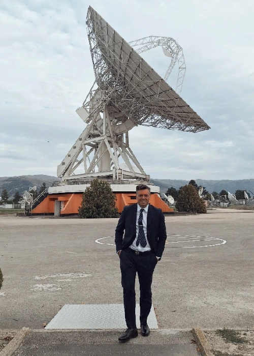 Un uomo in piedi, in giacca e cravatta, sullo sfondo un satellite