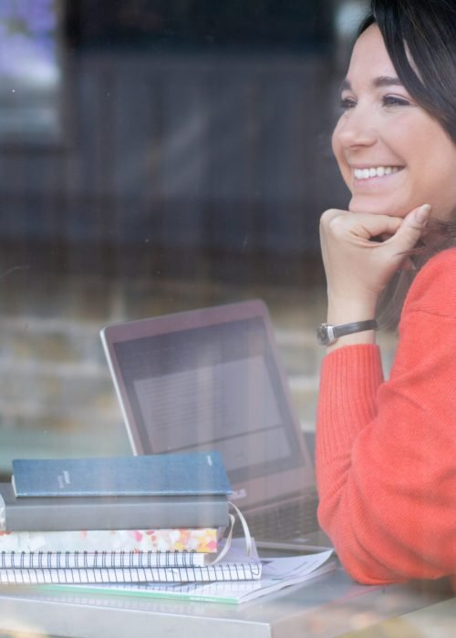 Una donna a mezzo busto che guarda fuori dalla finestra, ha davanti un laptop e il mento appoggiato al pugno chiuso, il gomito sul tavolo